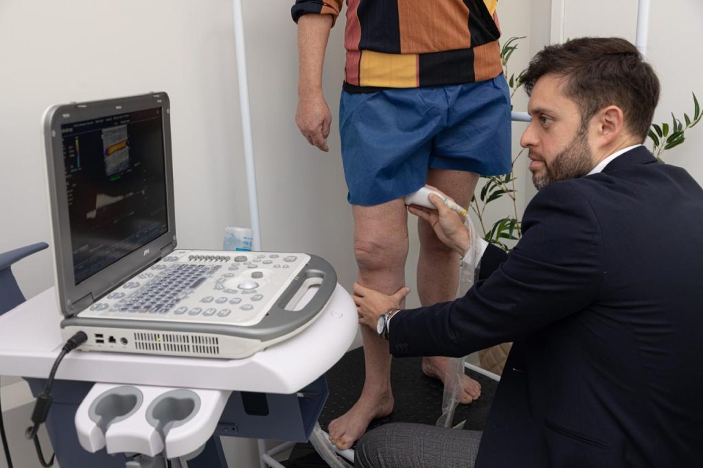 Um profissional médico usa um dispositivo de ultrassom para examinar o joelho de um paciente. O paciente está de pé e de bermuda, enquanto o médico segura a sonda de ultrassom contra o joelho e olha para a tela.