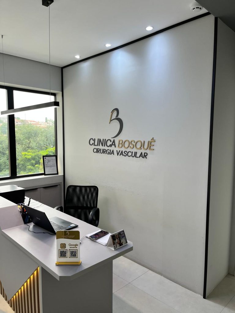 Área de recepção de clínica moderna com uma mesa, cadeira preta, laptop, certificado emoldurado, folhetos informativos e uma placa na parede onde se lê Clínica Bosque Cirurgia Vascular. Uma grande janela permite a entrada de luz natural.