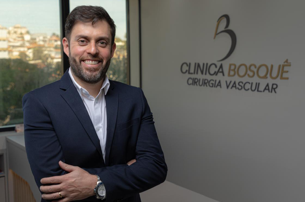 Um homem de terno sorri com os braços cruzados em um escritório, ao lado de uma placa que diz Clínica Bosqué Cirurgia Vascular. Uma janela com vista para edifícios está ao fundo.