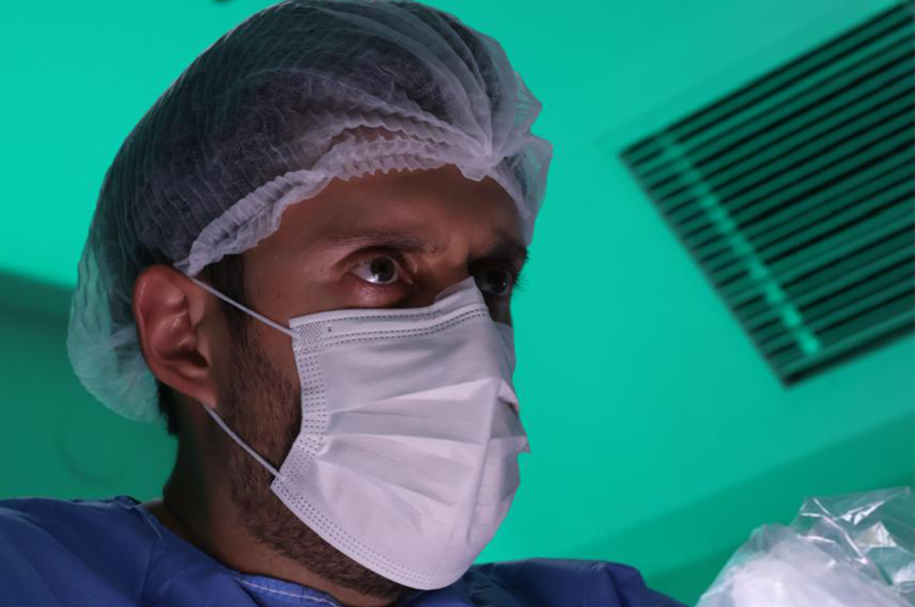 Um close-up de um profissional de saúde da Cirurgia Vascular usando touca cirúrgica, máscara facial e avental azul, em pé em um ambiente interno sob iluminação verde com um respiradouro visível ao fundo.
